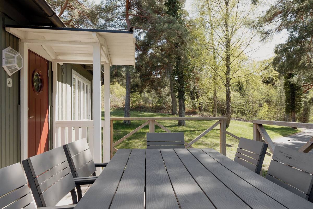 Une terrasse en bois avec une table de pique-nique et des bancs, entourée d’arbres et d’une pelouse.