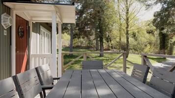 Una terraza de madera con una mesa de picnic y bancos, rodeada de árboles y un área cubierta de césped.