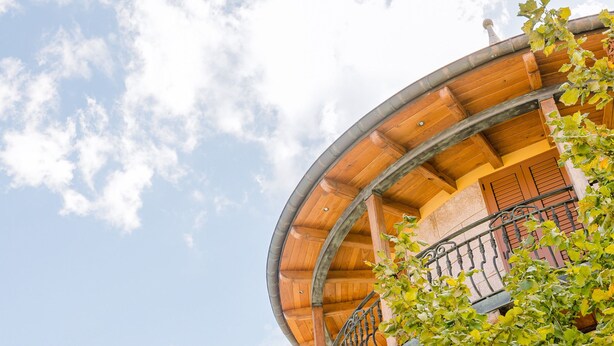 Ein runder, aus Holz gefertigter Balkon mit Metallgeländer, der auf ein Gebäude mit gelber Fassade blickt.
