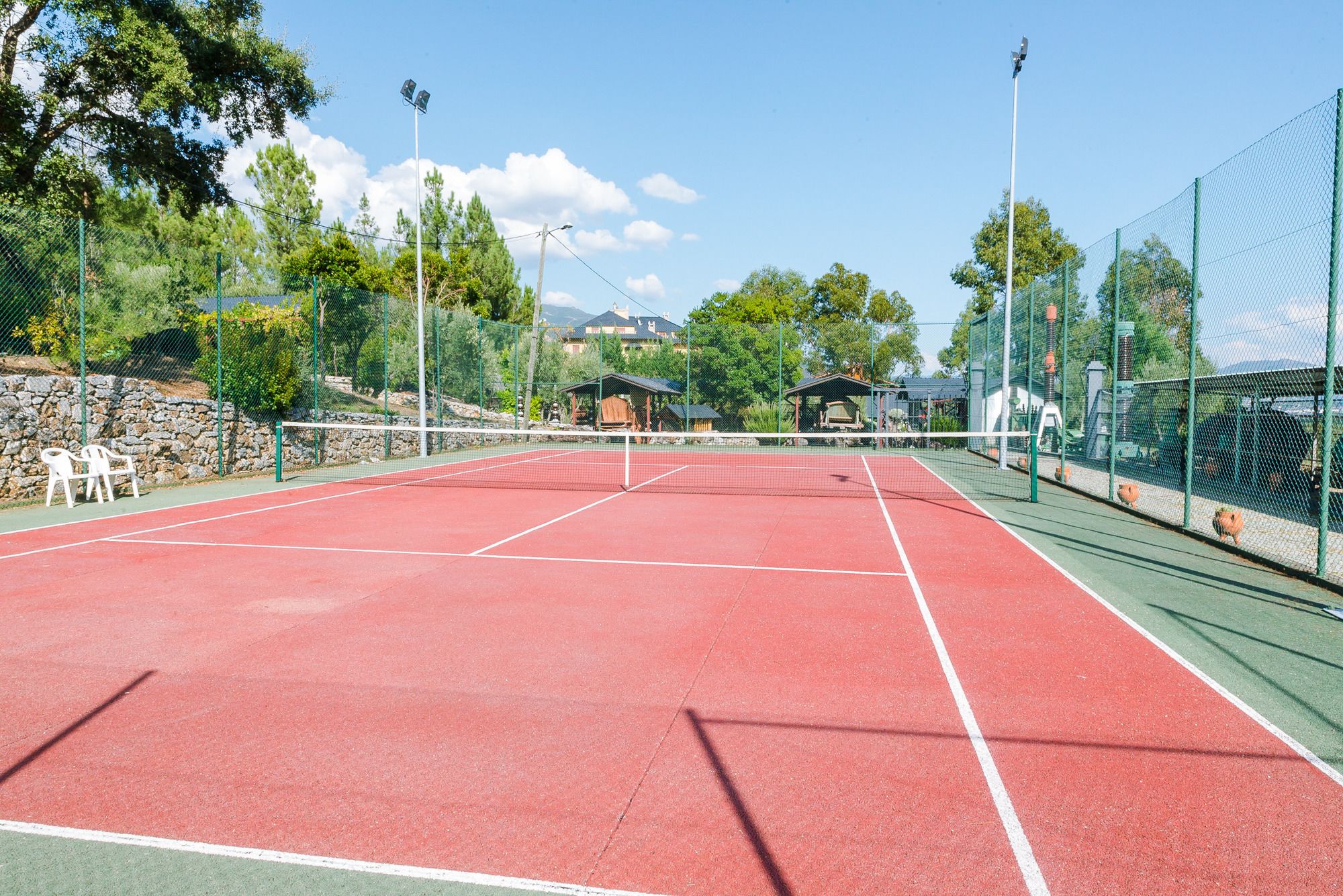 Una cancha de tenis al aire libre con red, rodeada de vallas verdes y árboles.