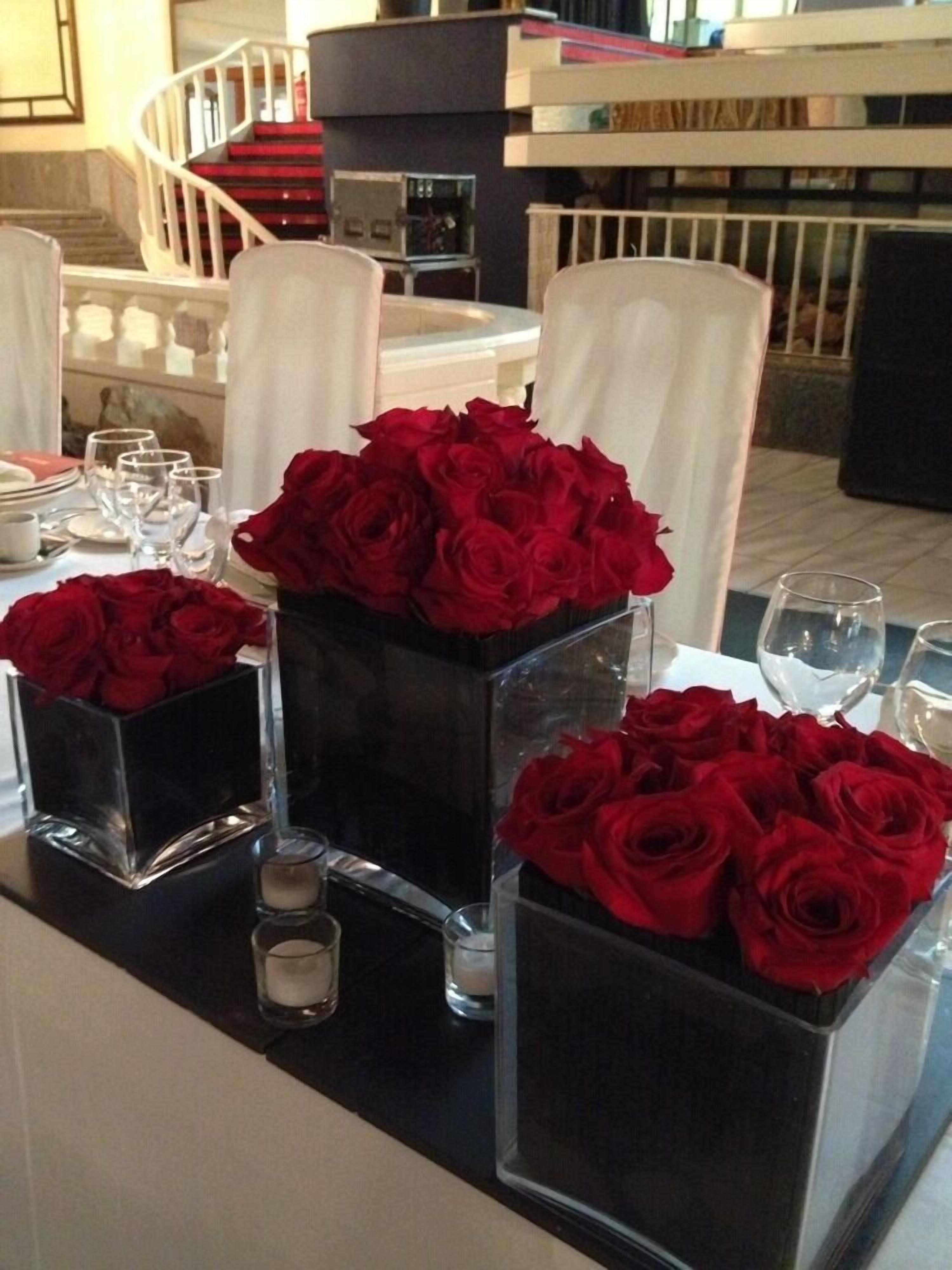 Una mesa de comedor con jarrones negros cuadrados que contienen rosas rojas, sillas blancas y una escalera al fondo.