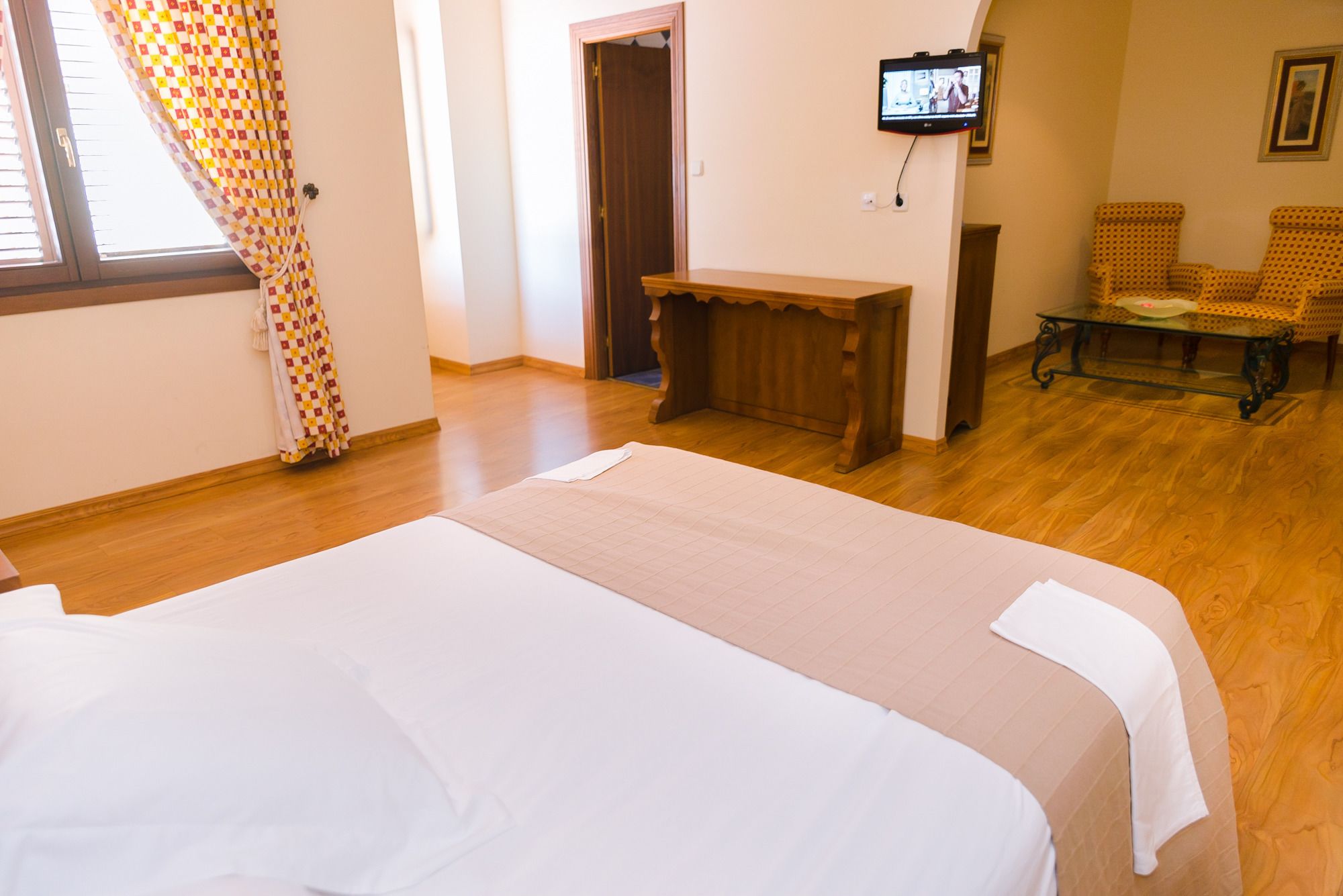 A hotel room with a large bed, a wooden bench, a television, and two patterned chairs.