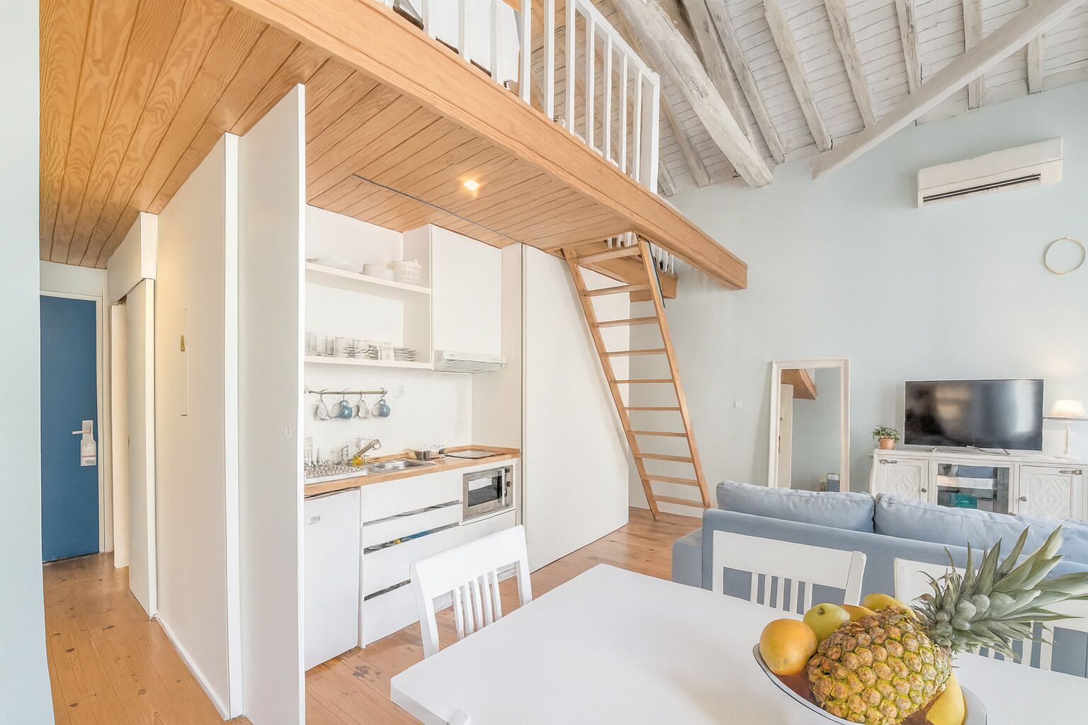 Una cocina moderna con armarios blancos, encimera de madera y horno empotrado. Hay una mesa de comedor con sillas y un frutero encima. Una escalera de madera lleva a una zona superior con televisor y asientos.