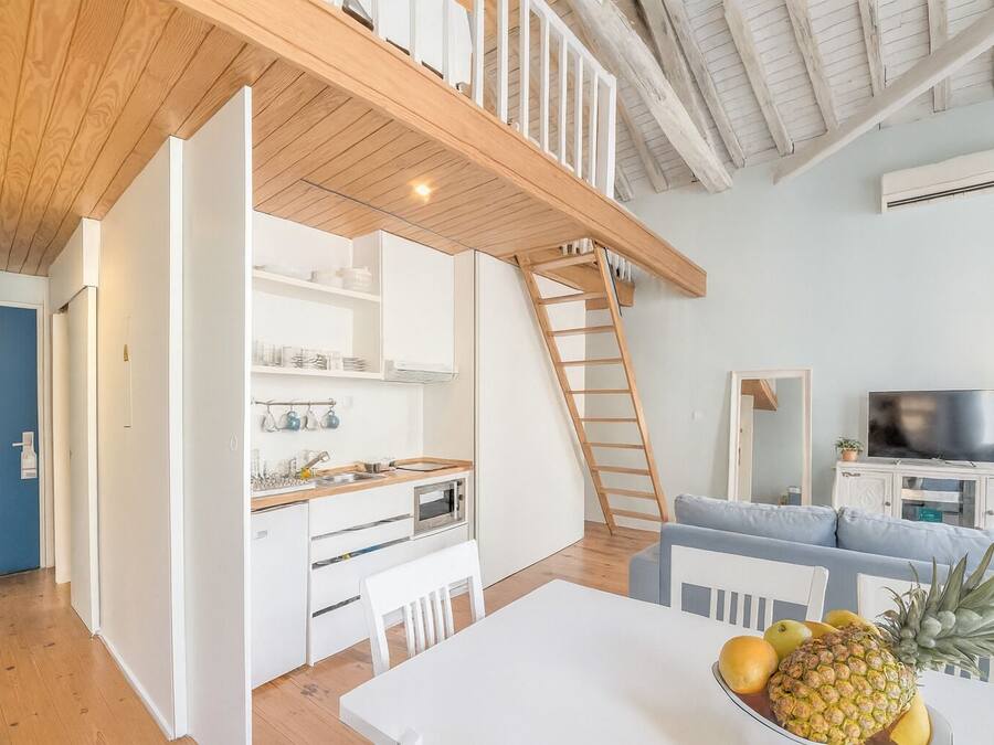 Una cocina moderna con armarios blancos, encimera de madera y horno empotrado. Hay una mesa de comedor con sillas y un frutero encima. Una escalera de madera lleva a una zona superior con televisor y asientos.