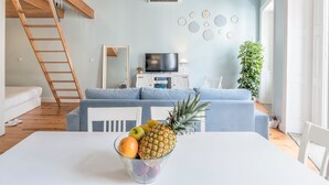 A modern living room with a white sofa, a dining table with fruit, a TV, and a staircase. - Look At Me - Serviced Lofts & Studios (Porto)