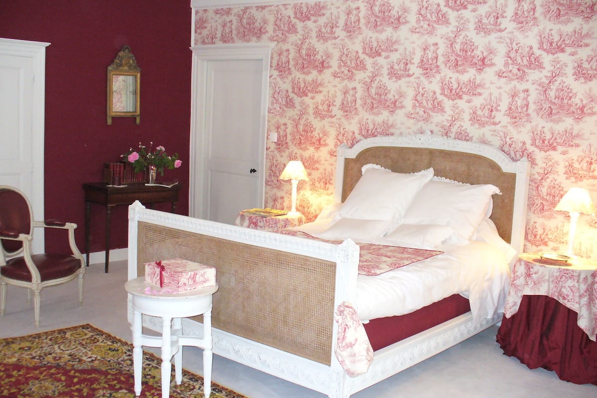 A bedroom with a four-poster bed, a small side table, a chair, and a patterned rug.