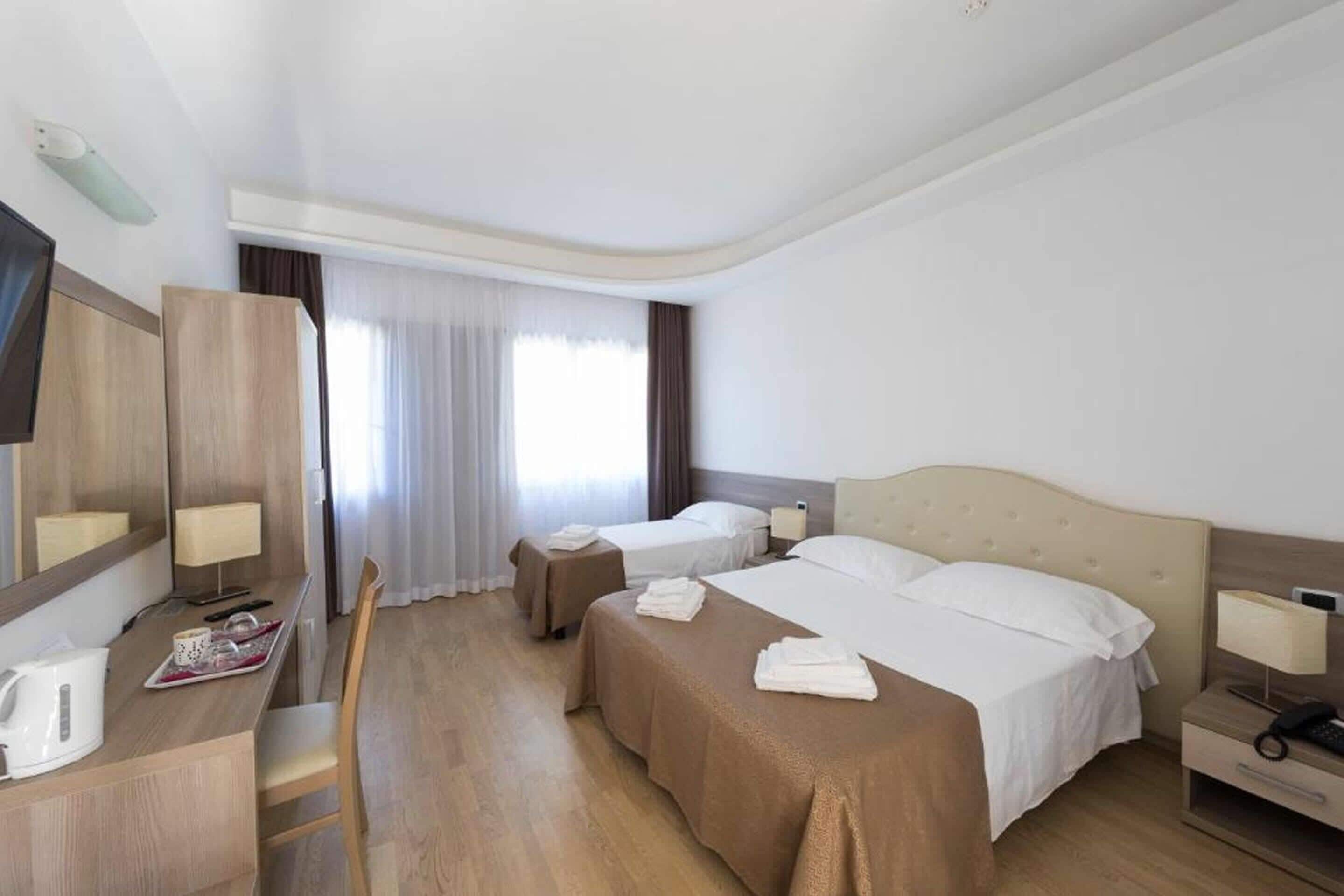 Habitación de hotel con dos camas, un escritorio con silla, un armario y una ventana con cortinas.