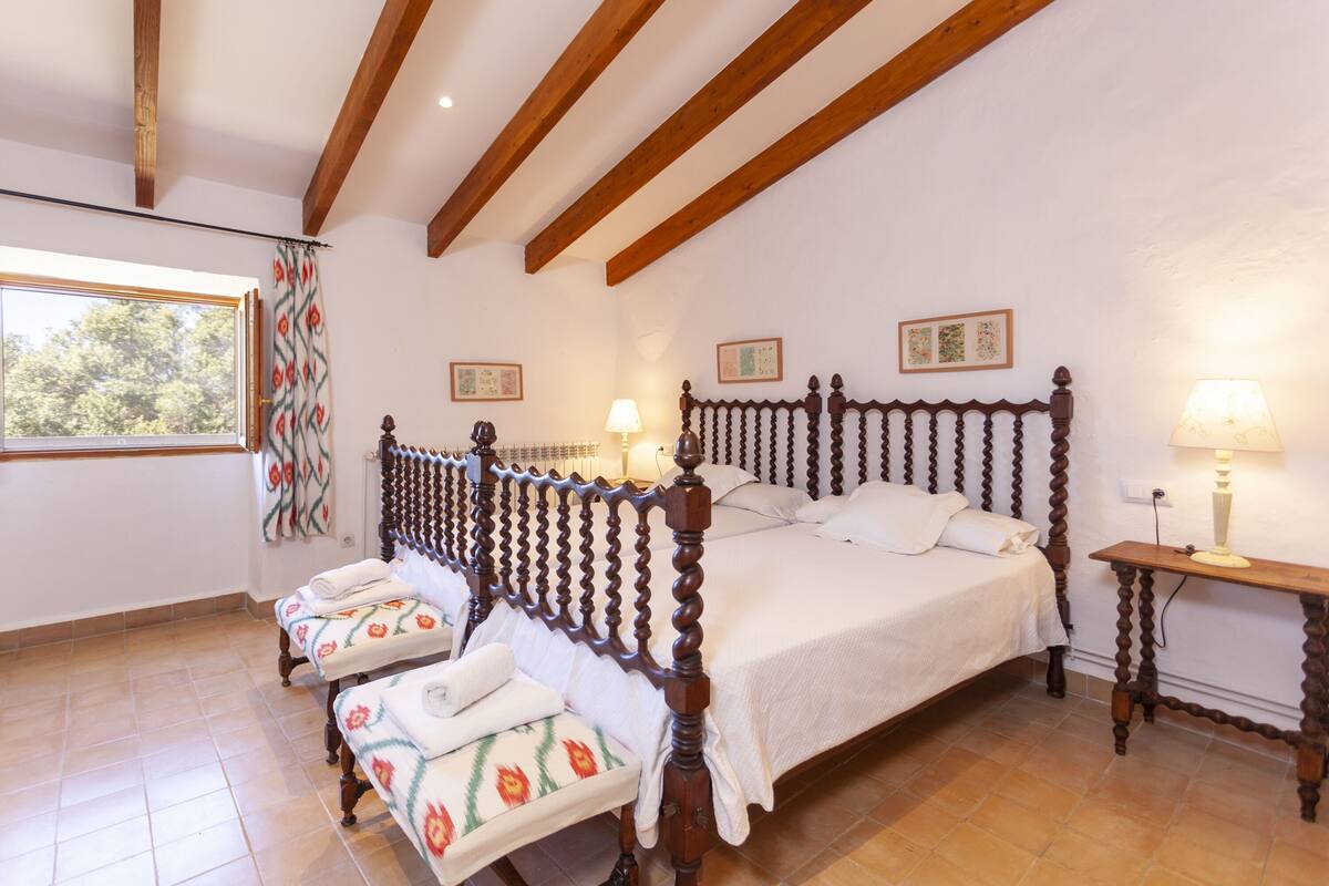 A bedroom with a wooden four-poster bed, a bench with floral cushions, a bedside table with a lamp, and a window with curtains.