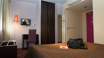 A hotel room with a bed, a desk, a chair, a TV, and a handbag on the bed.