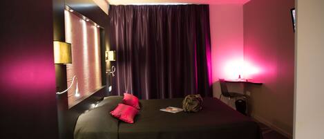 Una habitación de hotel con cama, almohadas, mesita de noche, lámpara y ventana con cortinas.
