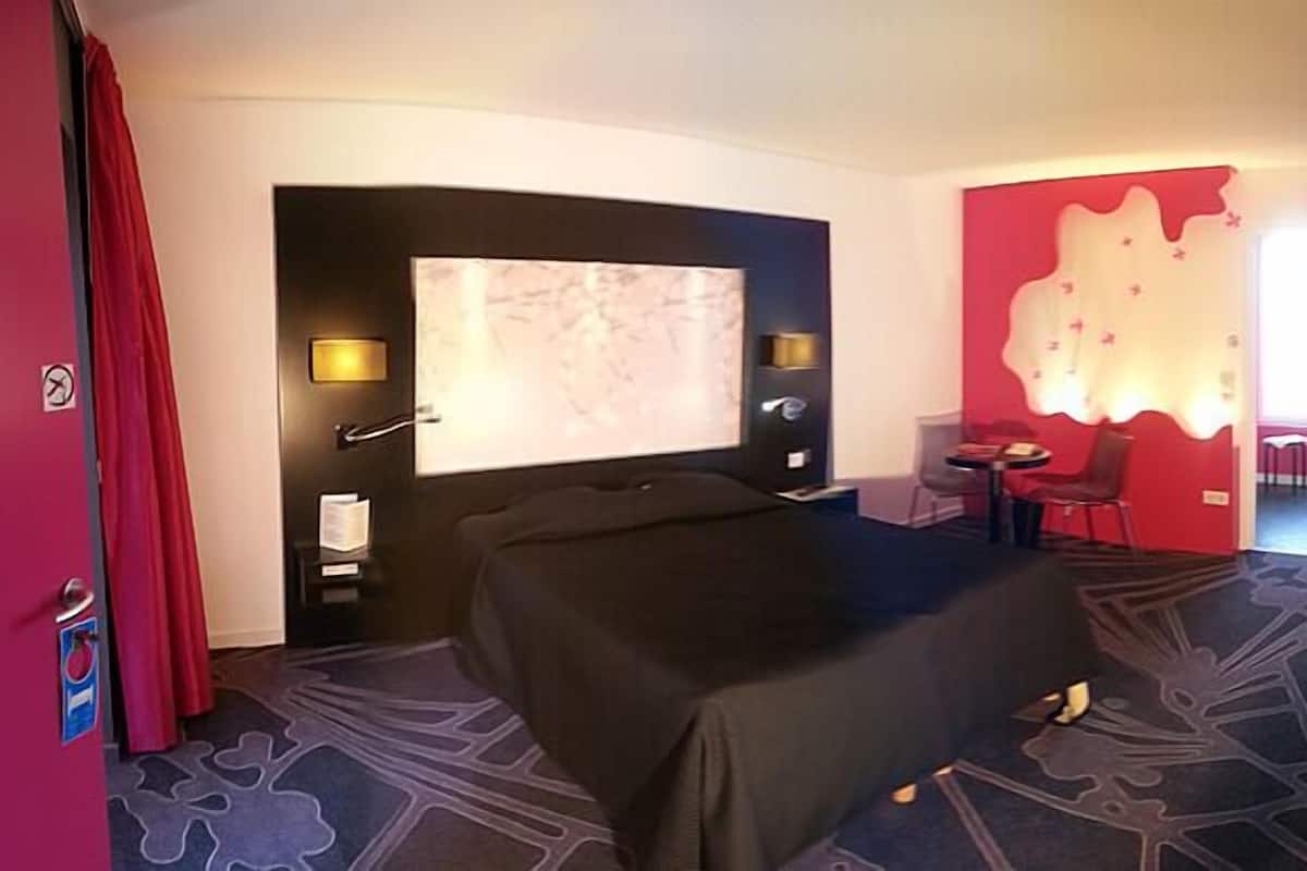 A hotel room with a bed, a desk, a chair, a small table, a red accent wall, and a large window.