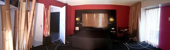 Habitación de hotel con cama curvada, un escritorio y una silla. Hay dos lámparas en la pared y una ventana con cortinas.