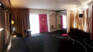 Habitación de hotel con cama, una mesita, una silla, un espejo y una ventana con cortinas.