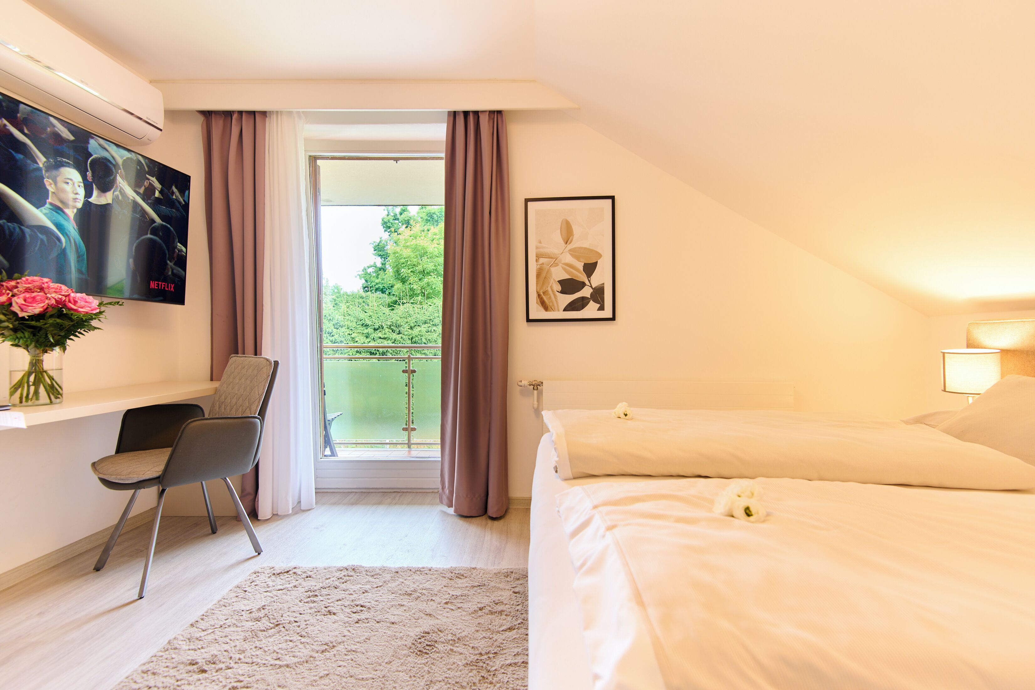 A hotel room with a bed, a desk with a chair, a TV mounted on the wall, and a window with curtains.