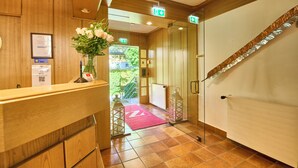 Una zona de recepción con mostrador de madera, un certificado enmarcado, un jarrón con flores y una alfombra roja que lleva a una puerta.