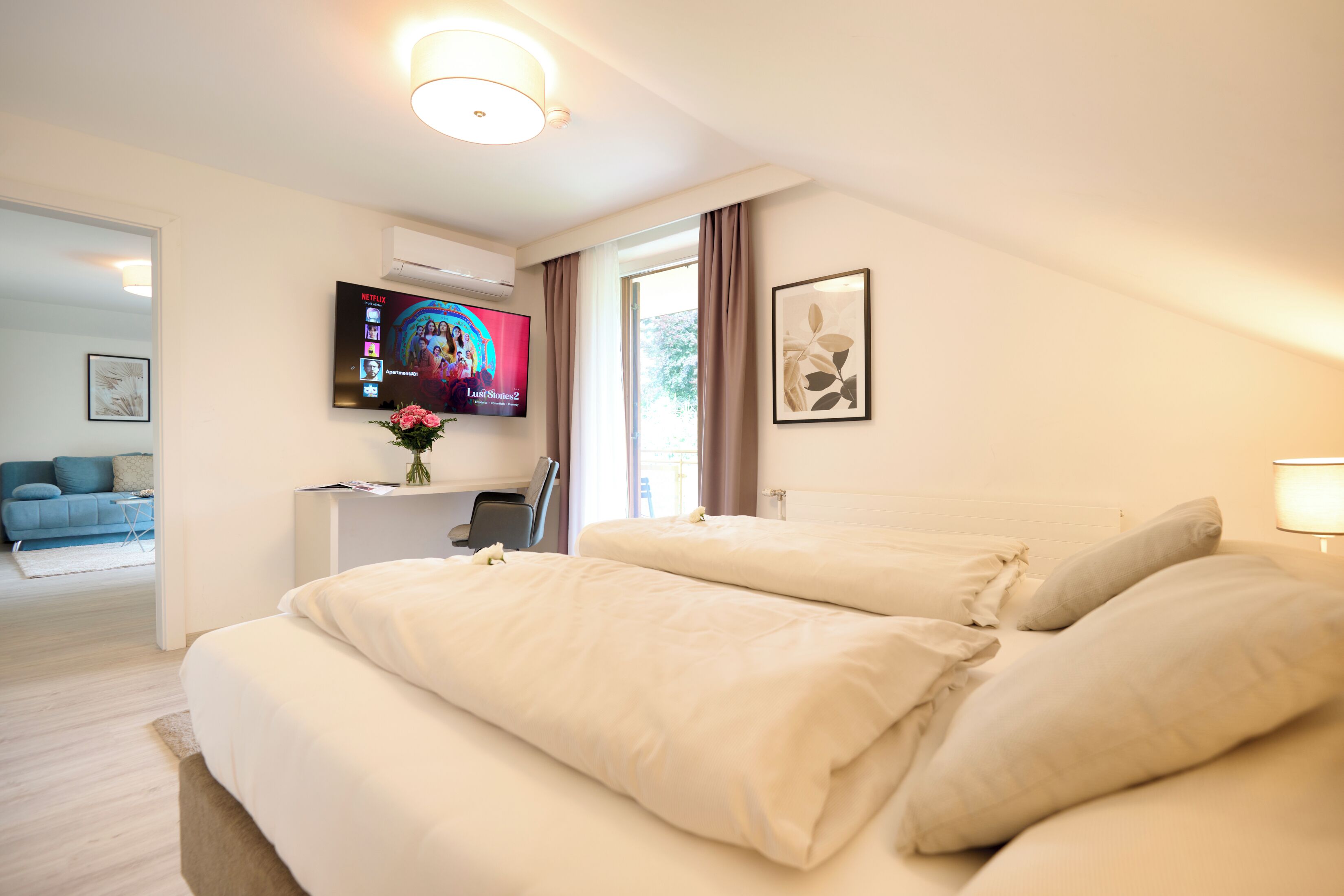 Habitación de hotel con una cama grande, un escritorio con computadora, un televisor montado en la pared y una ventana con cortinas.
