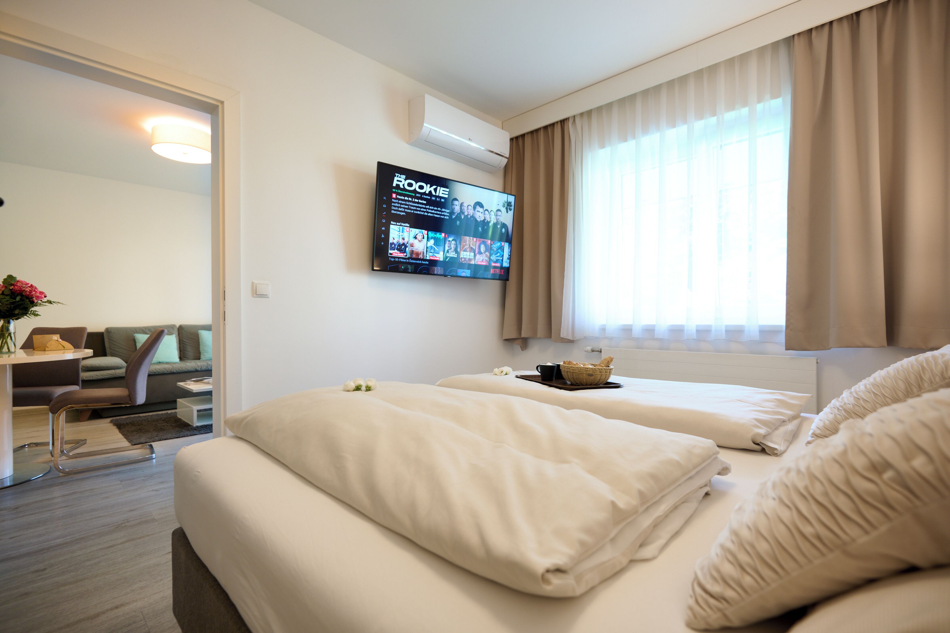 A hotel room with two beds, a flat-screen TV mounted on the wall, a window with curtains, and a small table with a basket.