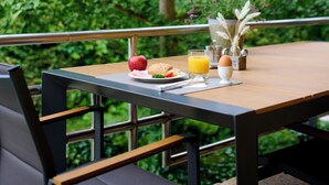 Una mesa servida con alimentos para el desayuno, como una manzana, un sándwich y jugo de naranja, con vista a una vegetación exuberante y un edificio al fondo.