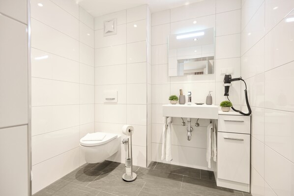A modern bathroom with white tiles, a toilet, a sink with a mirror, a hairdryer, and a towel rack. - Ferienwohnungen - Appartements Pension Zollner (Villach)