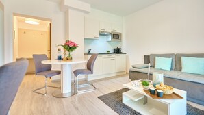 Una sala de estar moderna con un sofá gris, una mesa de comedor con dos sillas, una zona de cocina con armarios blancos y un microondas, y una alfombra gris en el suelo.