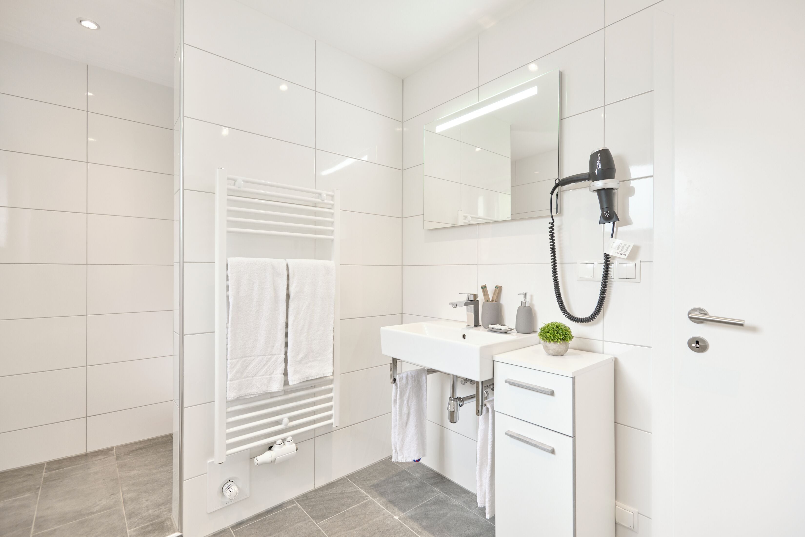 Une salle de bain moderne avec une douche à parois de verre, un lavabo blanc avec un miroir au-dessus, et un porte-serviettes avec des serviettes blanches.
