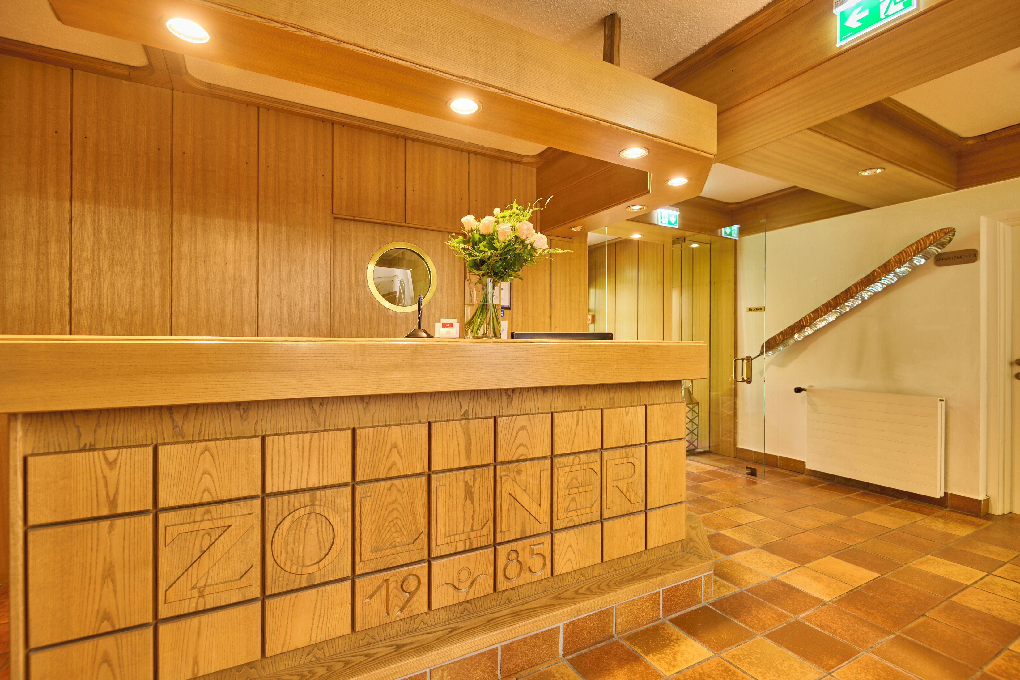 A wooden reception desk with a mirror and a vase of flowers. The reception area has a tiled floor and a wooden ceiling with recessed lighting.