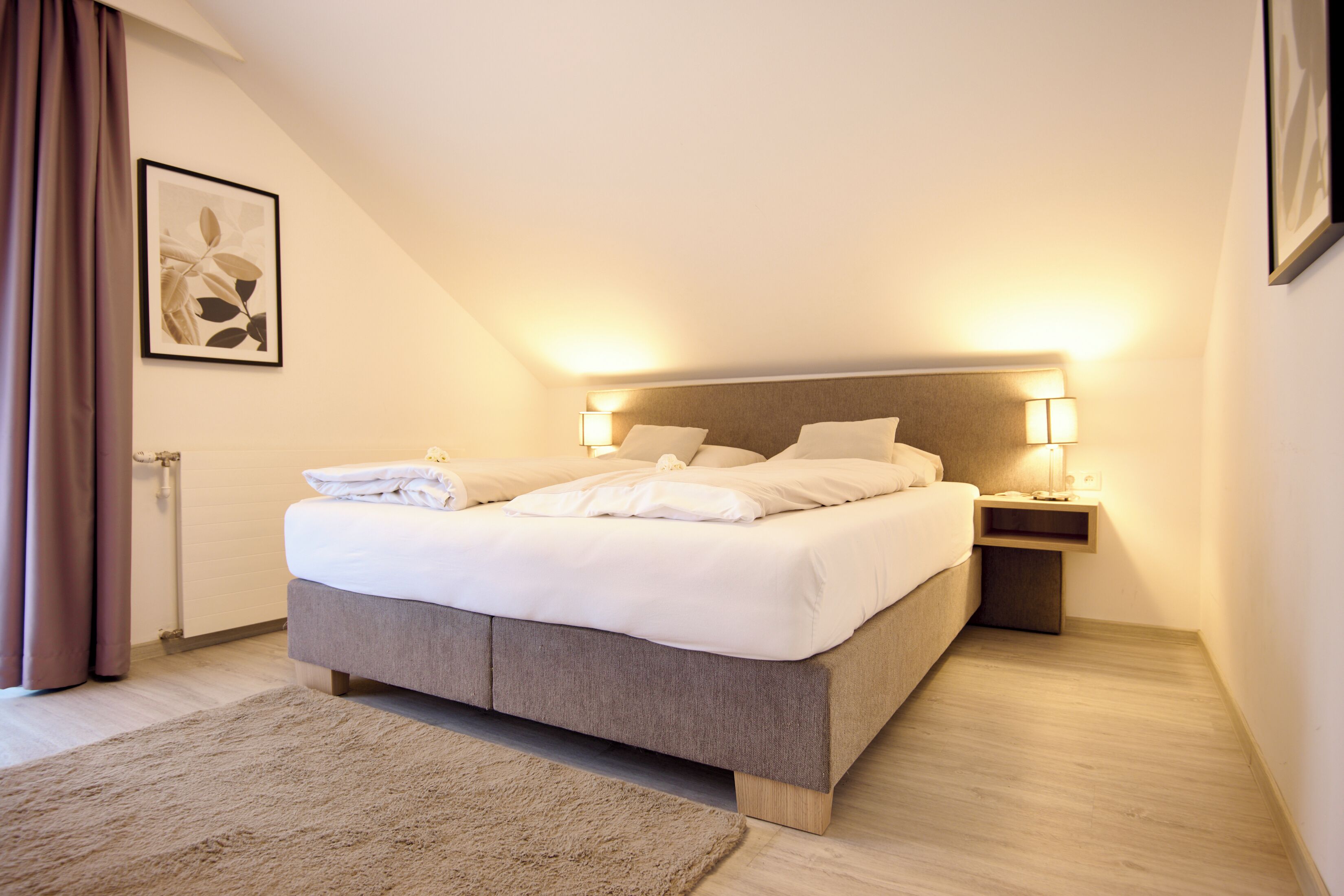 Ein modernes Schlafzimmer mit einem großen Bett, zwei Nachttischen mit Lampen, einem gerahmten Bild an der Wand und einem Fenster mit Vorhängen.