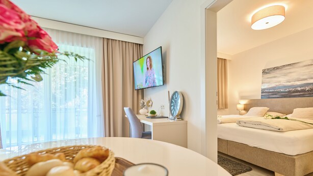 Habitación de hotel con cama, televisor, zona de comedor con mesa y sillas, y ventana con cortinas.