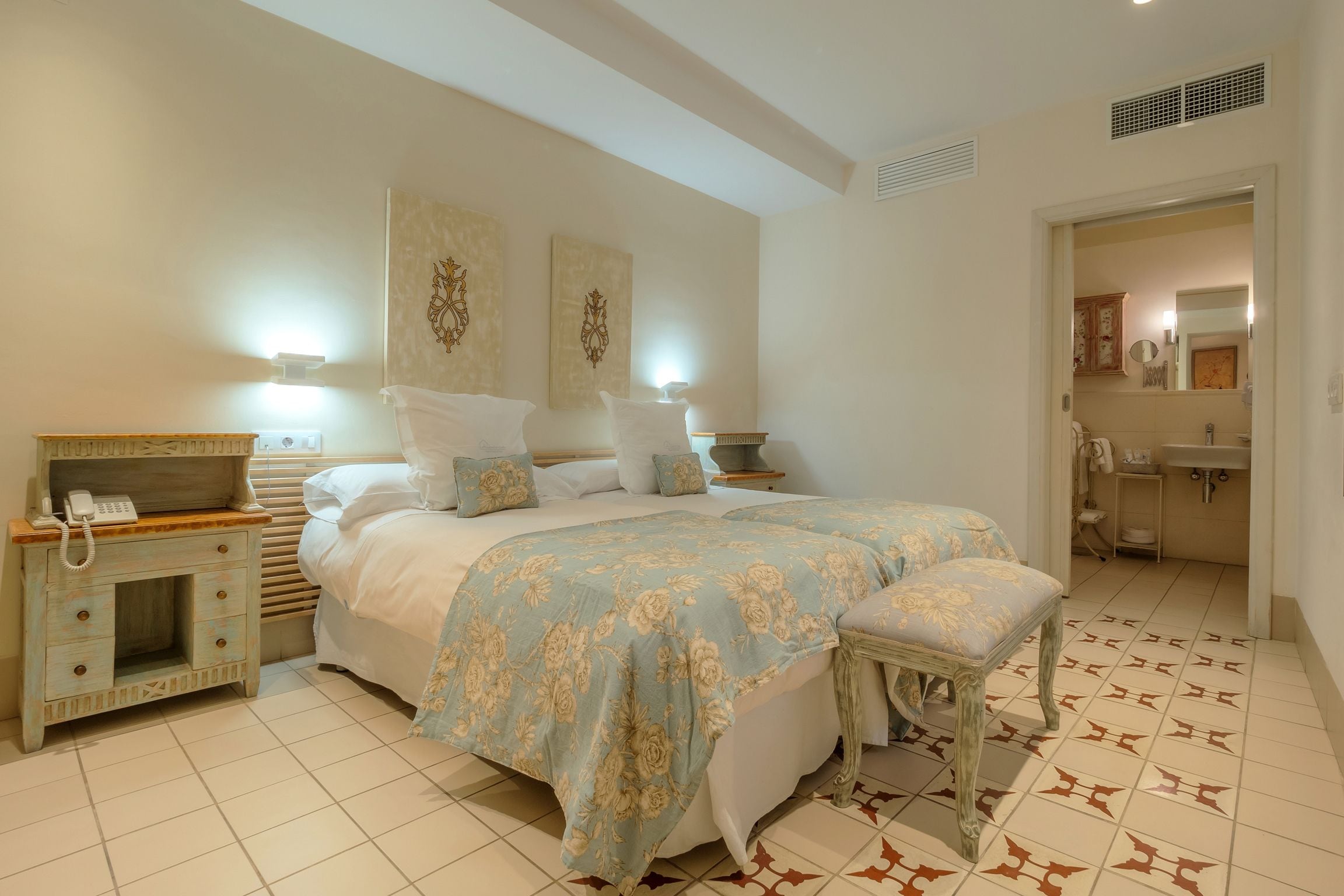 Ein Schlafzimmer mit einem großen Bett, Nachttischen, einer Bank und einem durch eine offene Tür einsehbaren Badezimmer.