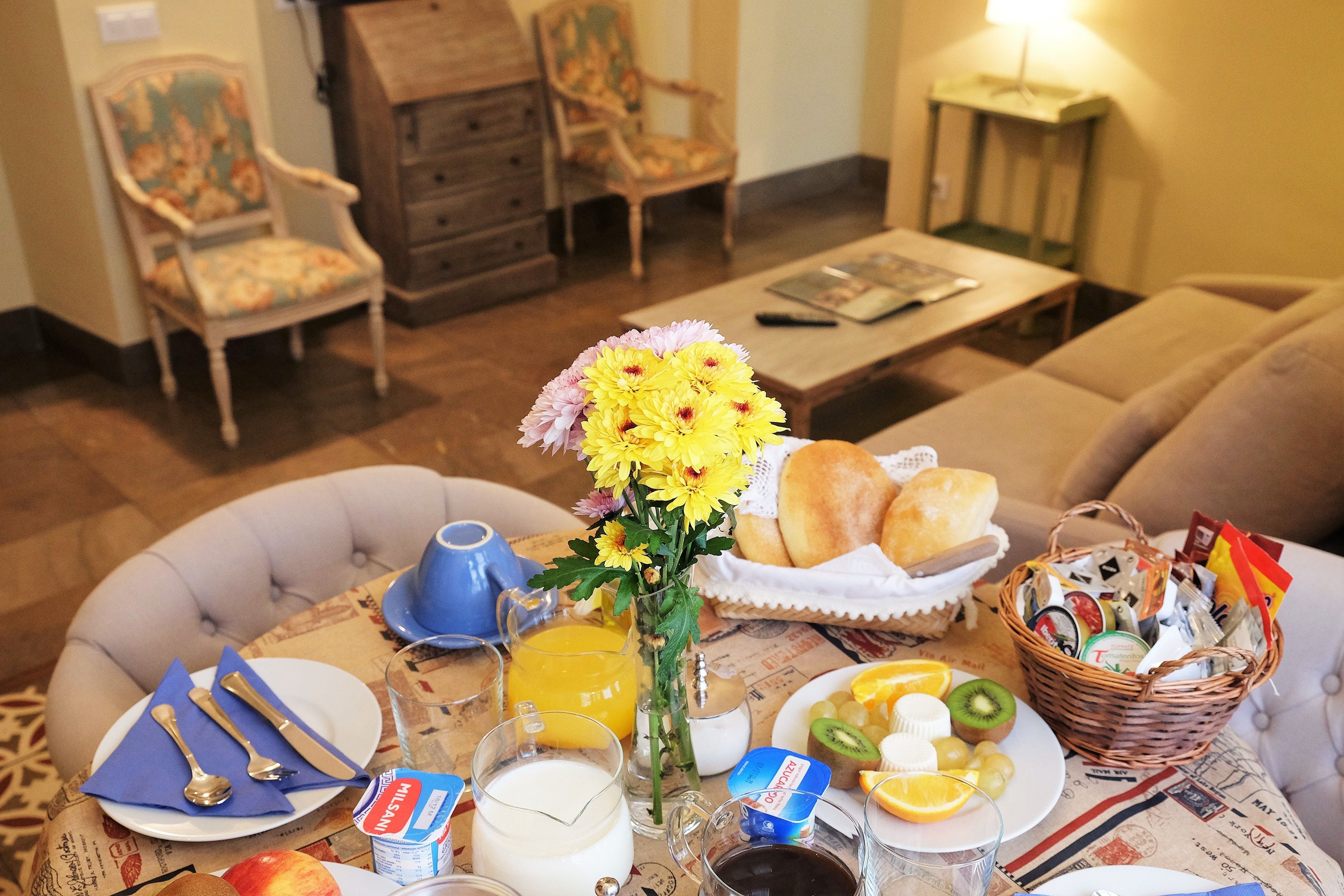 A breakfast setting with a table set for two, including plates, cutlery, a vase of flowers, a basket of bread, and various breakfast items.