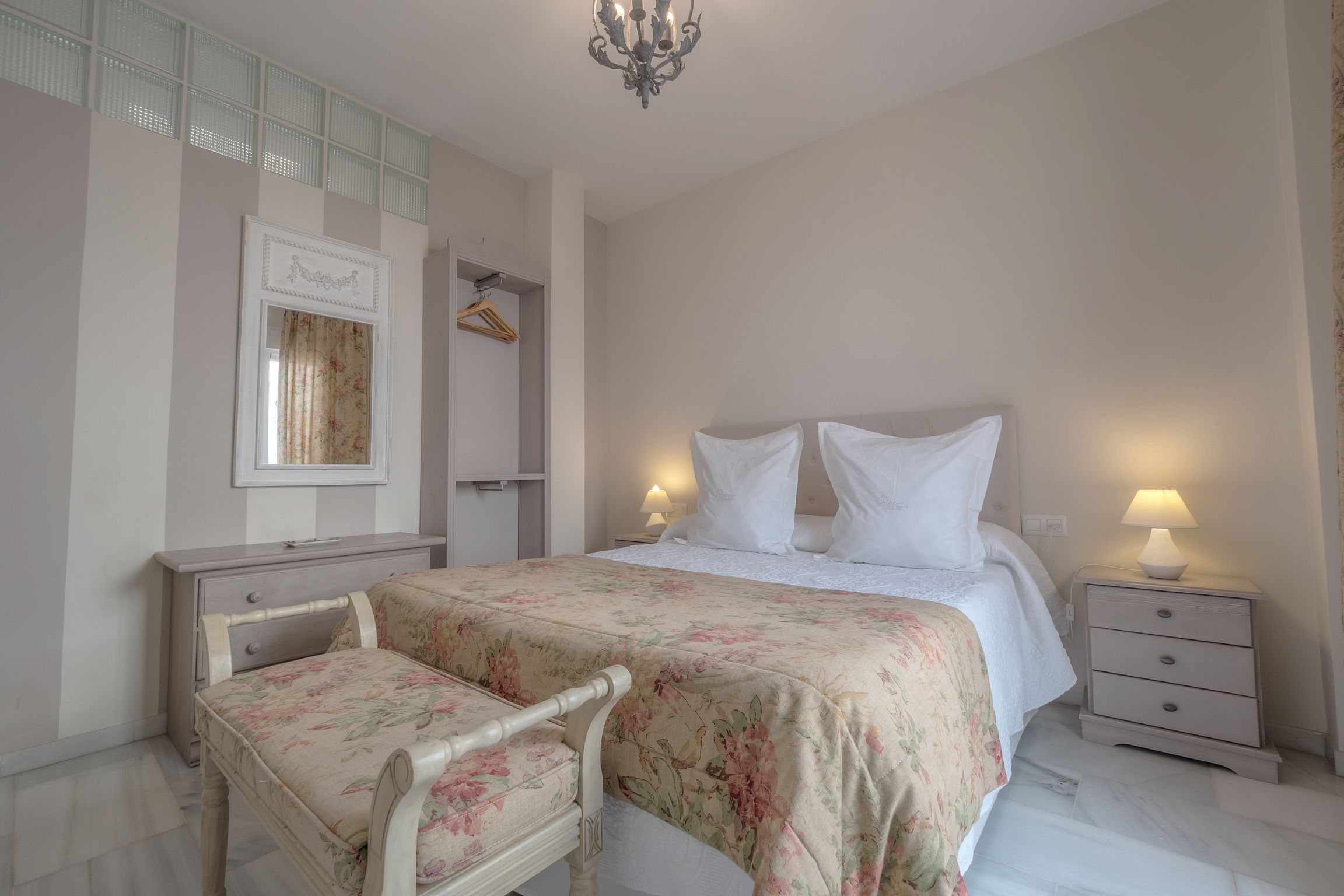 A bedroom with a bed, bedside tables, a chair, a wardrobe, and a chandelier.