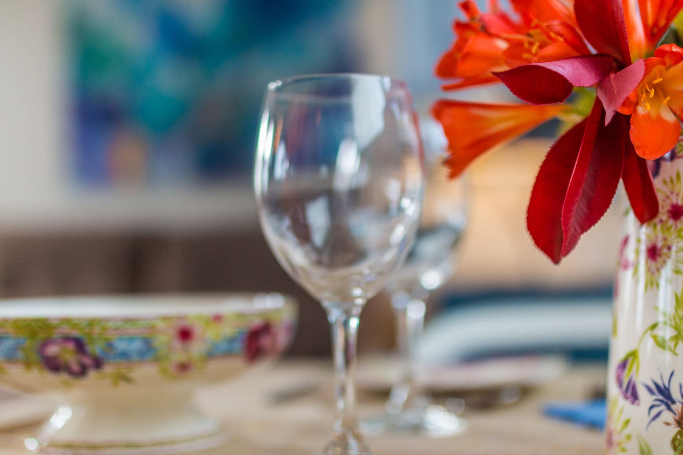 A dining table with a floral-patterned bowl, a clear wine glass, and a vase with red flowers.
