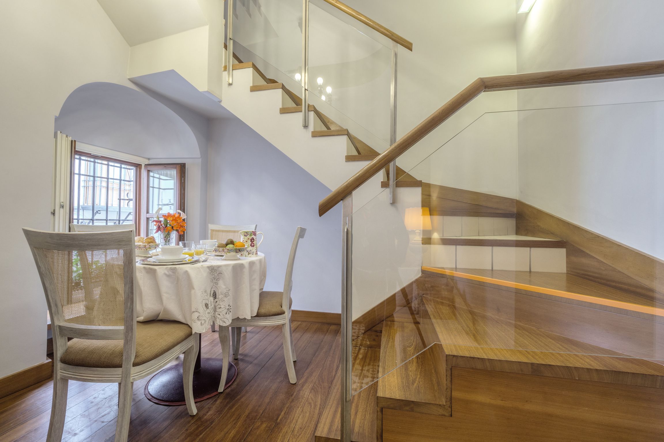 A dining area with a table set for four, a chair, and a staircase with wooden steps and glass railings.