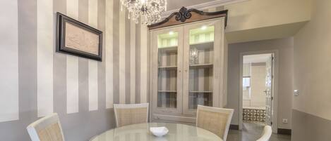 A dining area with a round glass table, four chairs, a chandelier, and a cabinet with glass doors.
