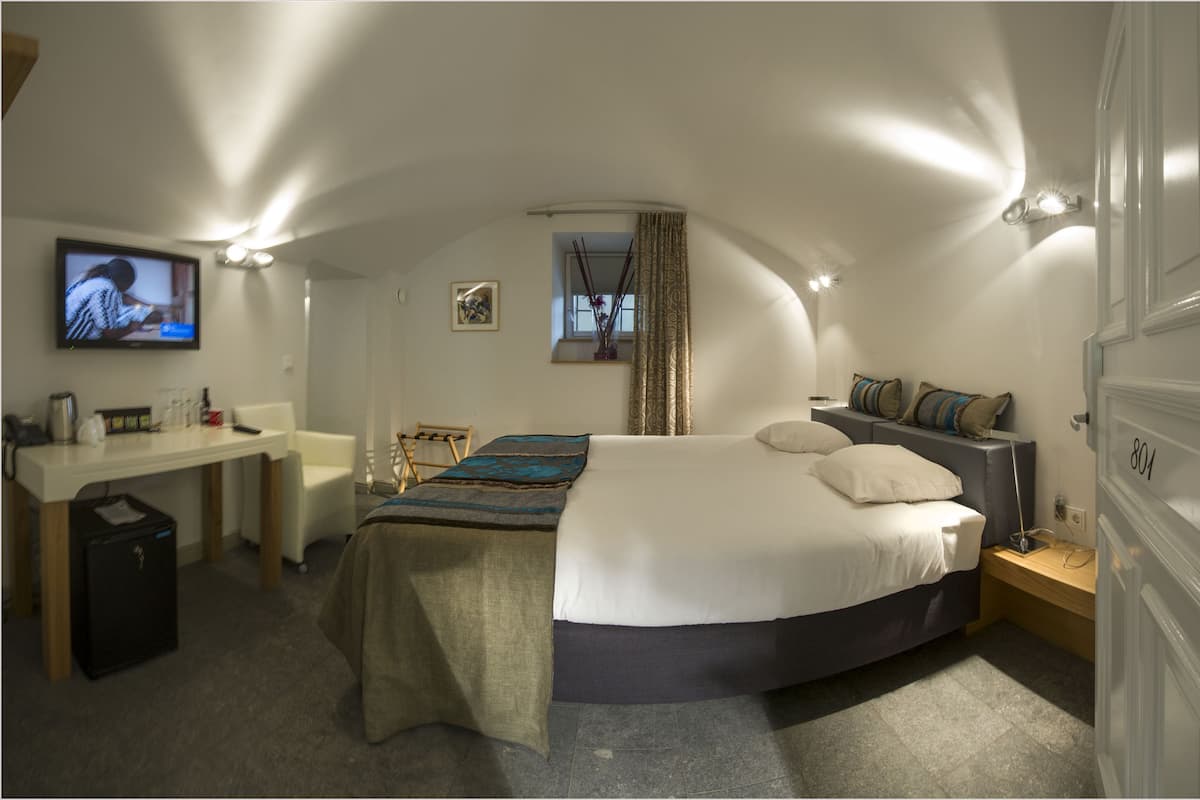 A bedroom with a bed, bedside table, TV, desk, chair, and a small window with curtains.