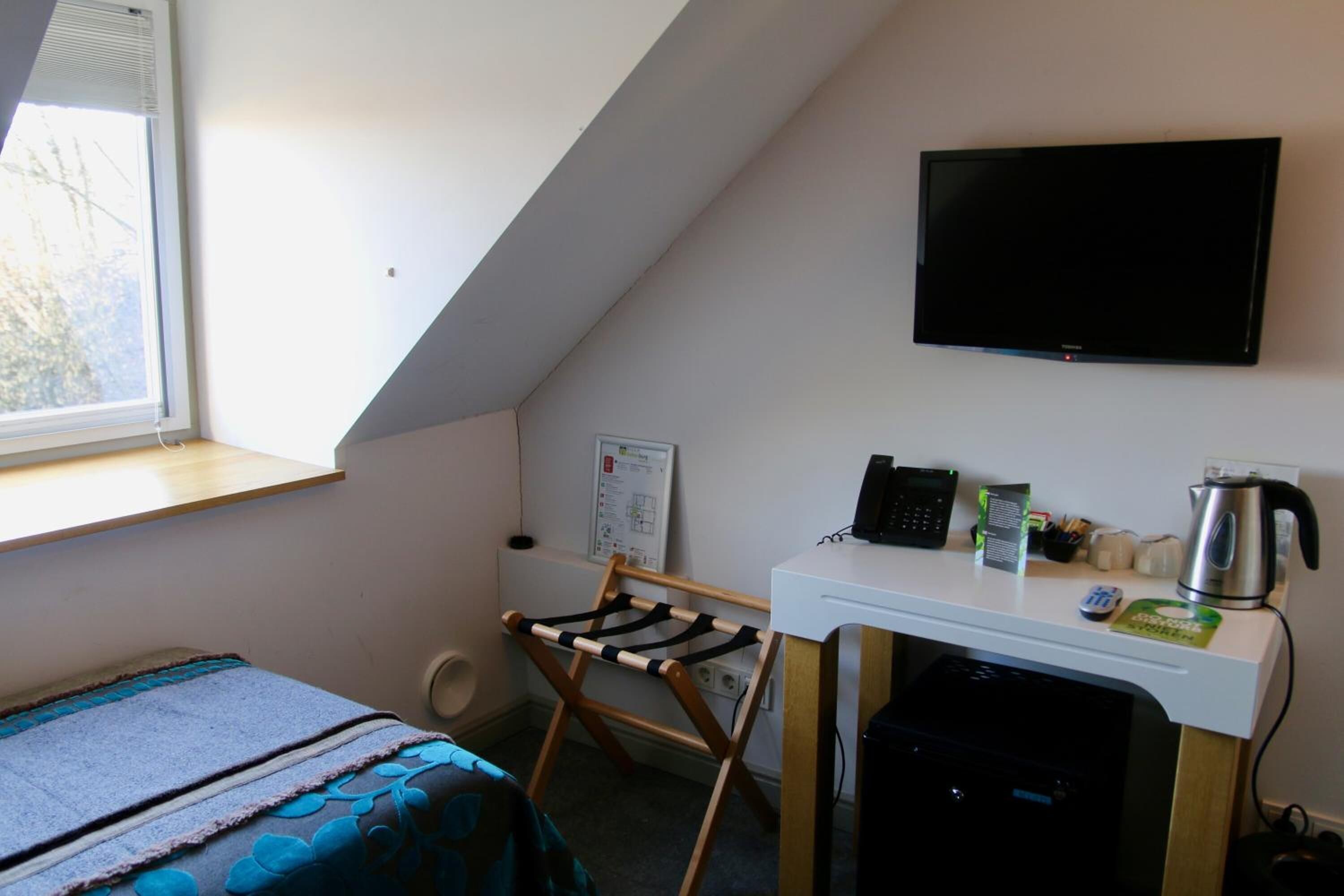 Une petite pièce bien éclairée, équipée d’un lit, d’un bureau avec un ordinateur et une bouilloire, d’un téléviseur fixé au mur et d’une fenêtre laissant entrer la lumière naturelle.
