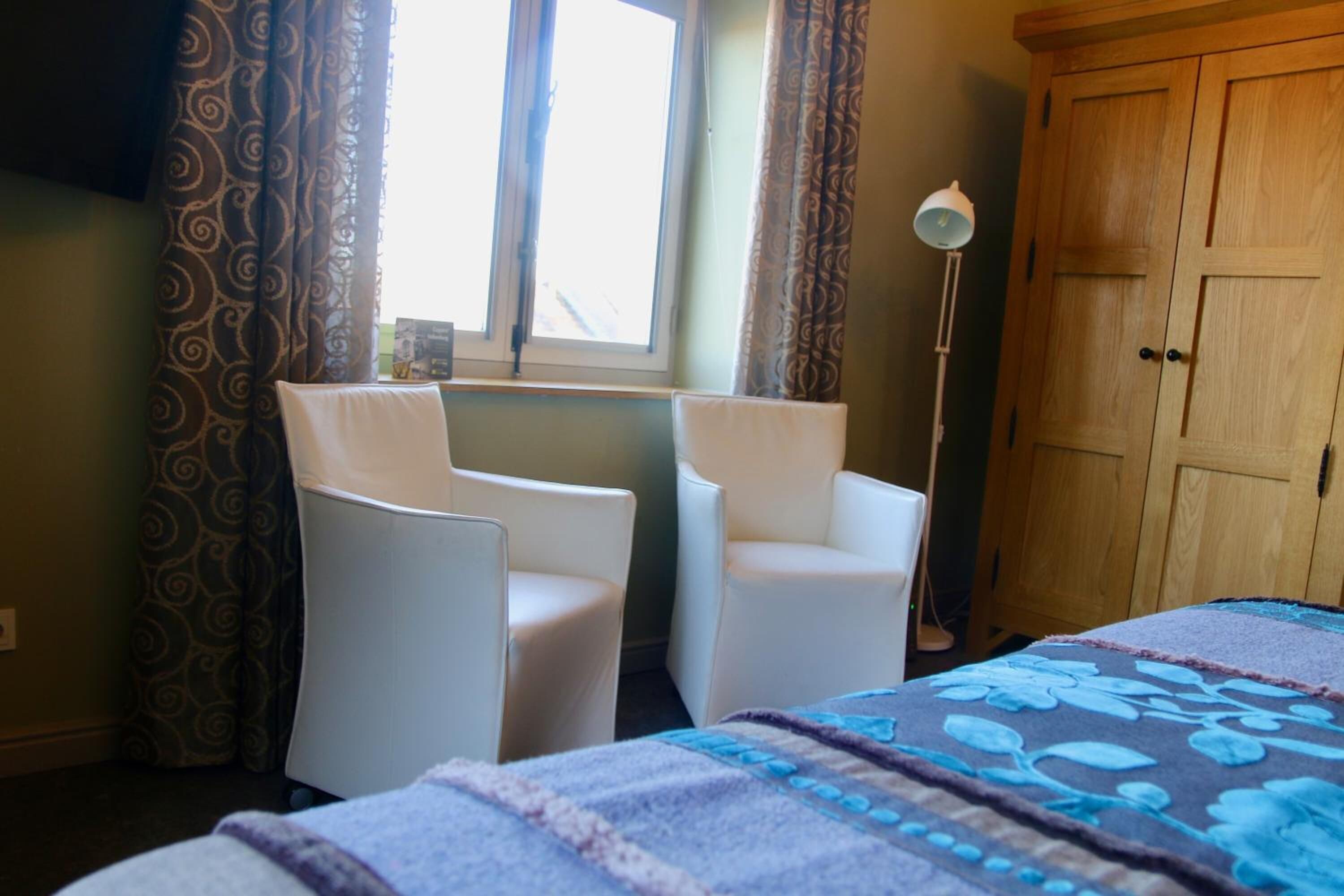 Een kamer met twee witte stoelen, een bed met een bloemenprintdeken, een houten garderobe en een vloerlamp.