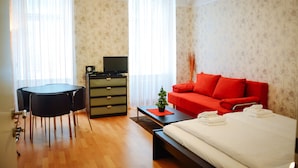 A hotel room with a bed, a red sofa, a dining table with chairs, a TV, and a small plant. - JL Apartments (Vienna)