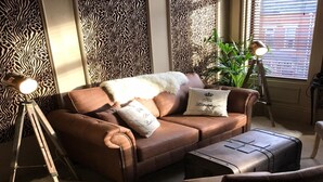A cozy living room with a brown leather sofa, two matching armchairs, a small ottoman, and decorative pillows. There are two floor lamps and a potted plant. - Kingsmere Guest House (South Shields)