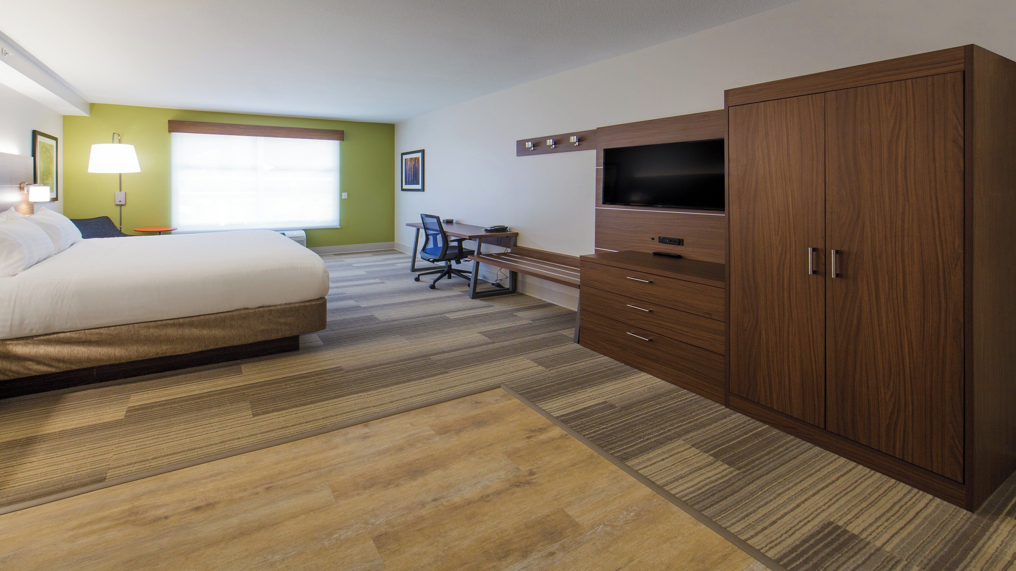 A hotel room with a bed, desk, chair, TV, wardrobe, and a window with blinds.