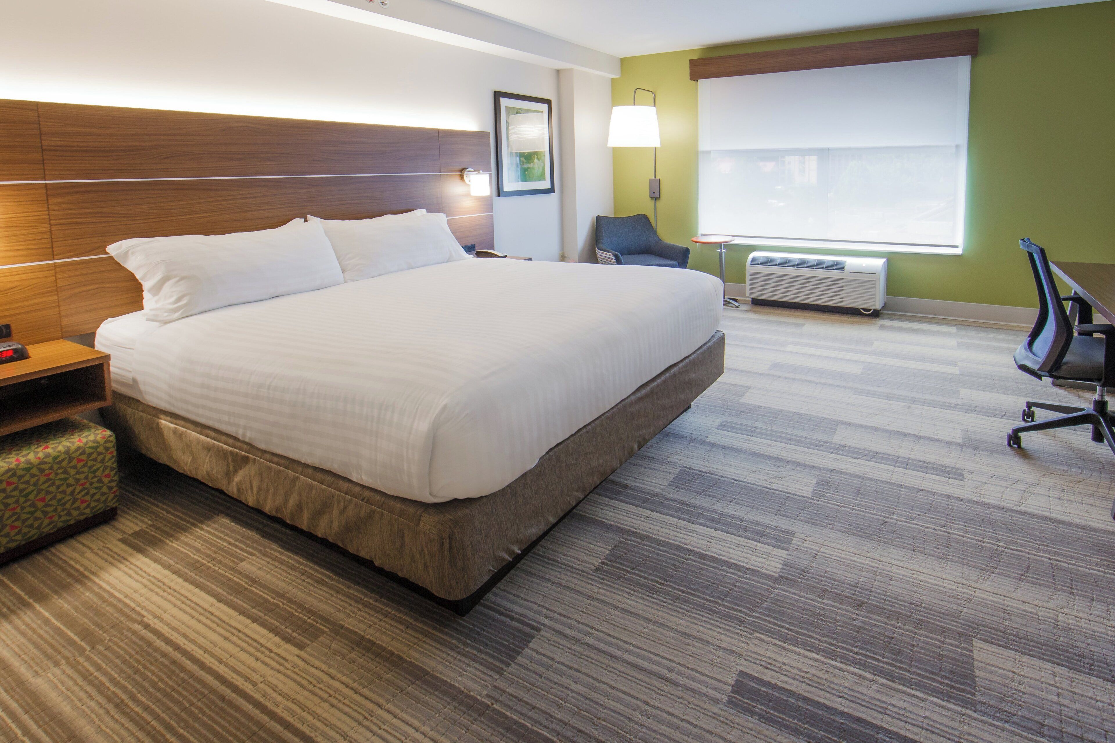 Ein Hotelzimmer mit einem großen Bett, einem Nachttisch, einem Sessel, einem Schreibtisch, einer Lampe, einem Fenster und einer Klimaanlage.