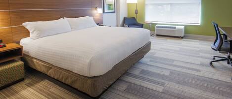 A hotel room with a large bed, a bedside table, a chair, a desk, a lamp, a window, and an air conditioning unit.