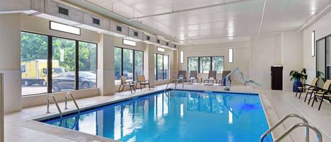 Una piscina interior con ventanales amplios, suelo de baldosas y algunas sillas de descanso.