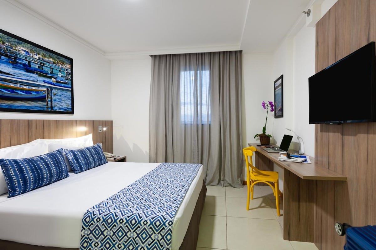 A hotel room with a bed, a desk, a chair, a television, and a window with curtains.