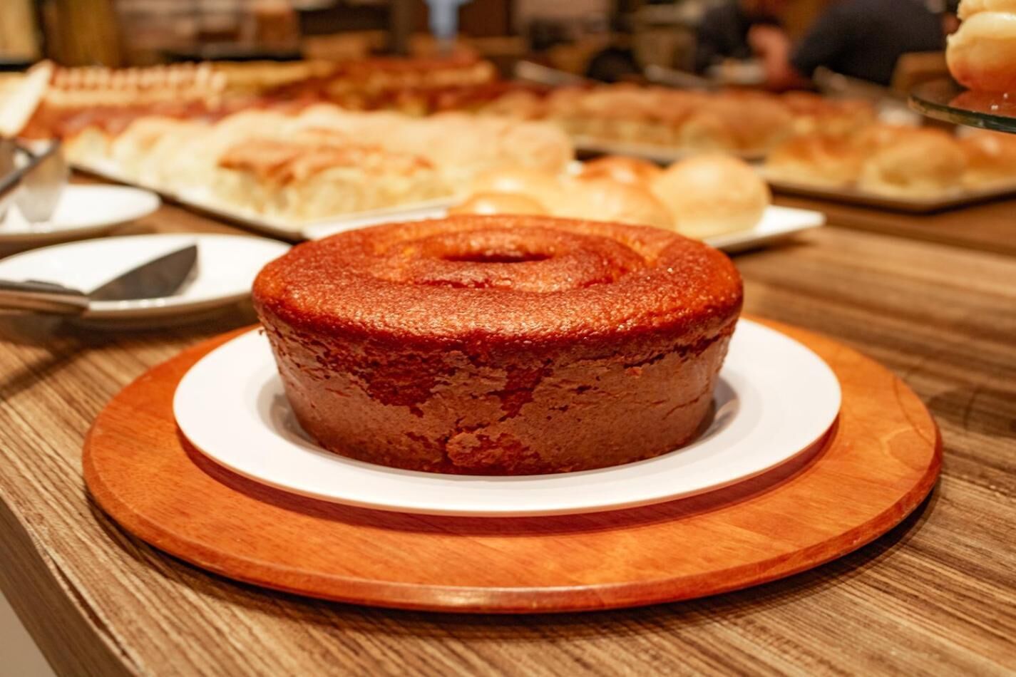 A round cake on a wooden board with a white plate underneath, placed on a wooden table.