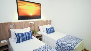 A hotel room with two beds, a wooden headboard, a wall-mounted TV displaying a sunset, and a small bedside table with a phone and a plant.
