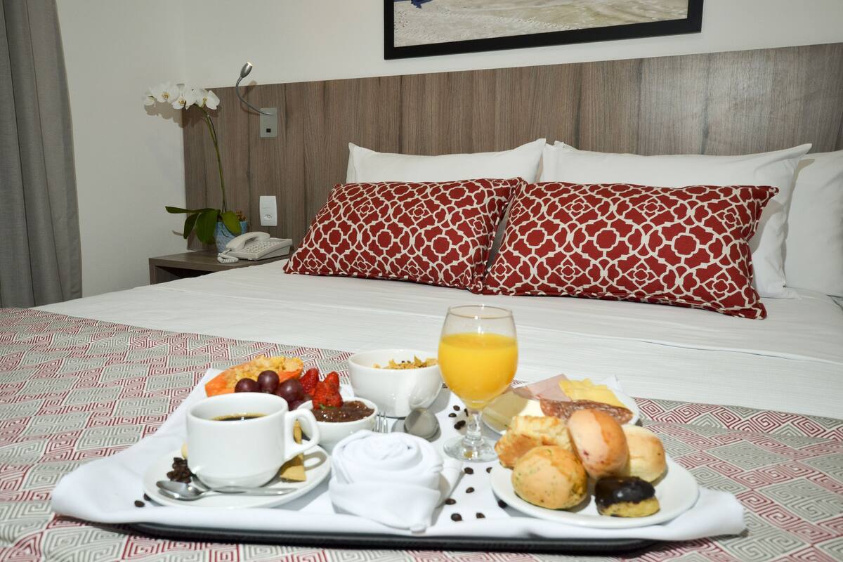 Un lit bien fait, agrémenté d’oreillers blancs et rouges à motifs, une table de chevet avec un téléphone et un vase de fleurs blanches, et un plateau de petit-déjeuner avec du café, des céréales, des fruits et des viennoiseries.