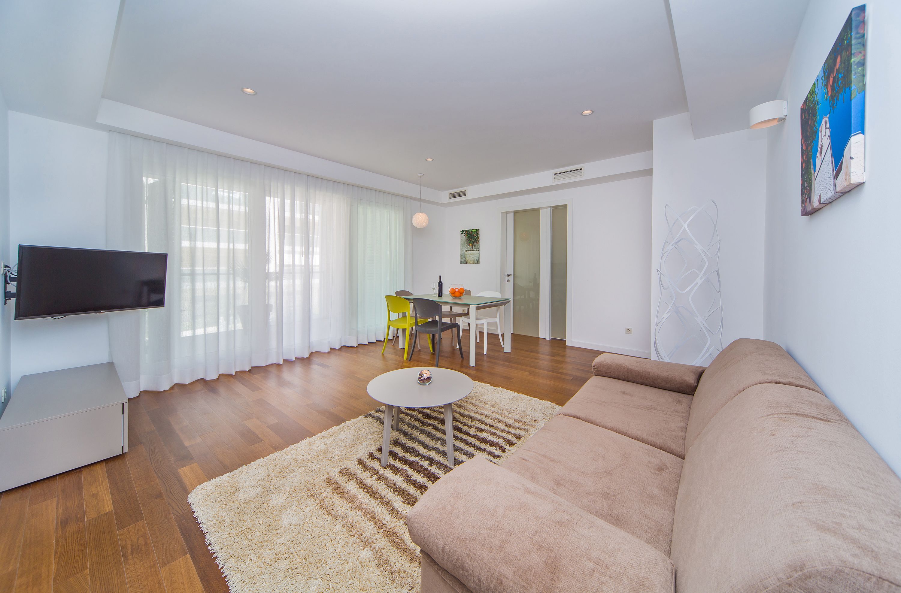Uma sala de estar moderna com uma televisão de tela plana, um sofá bege, uma mesa de centro redonda e uma área de jantar com mesa e cadeiras.