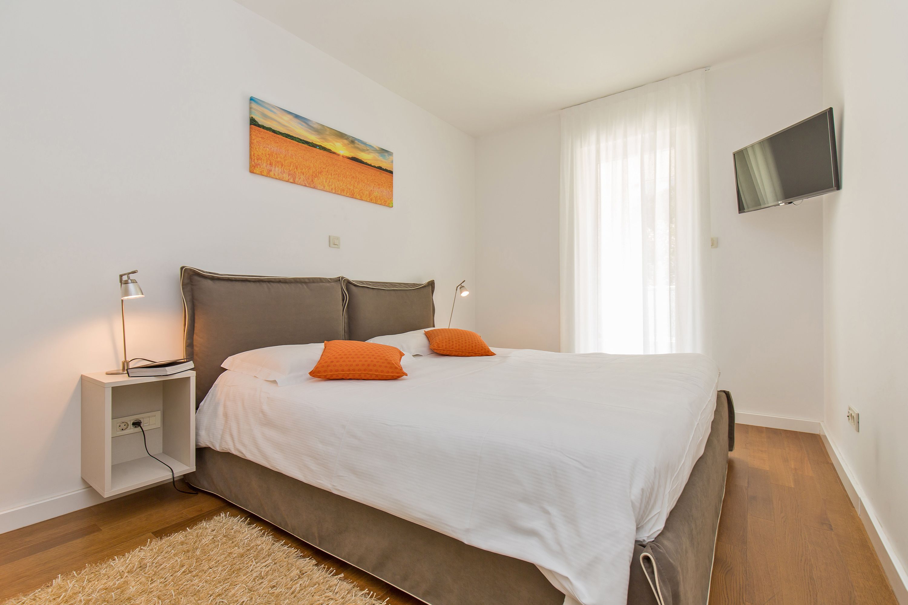 A bedroom with a bed, two bedside lamps, a painting on the wall, and a TV mounted on the wall.
