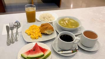Una mesa de desayuno con platos de huevos revueltos, un croissant, arroz, sopa y una rebanada de sandía.